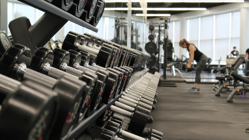An image of a gym with dumbbells in front and a woman in the back